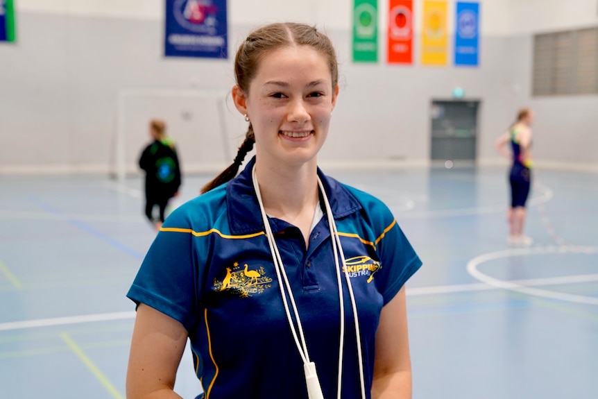 A young woman with two light brown braids and a jump rope around her neck stands in a gym smiling.