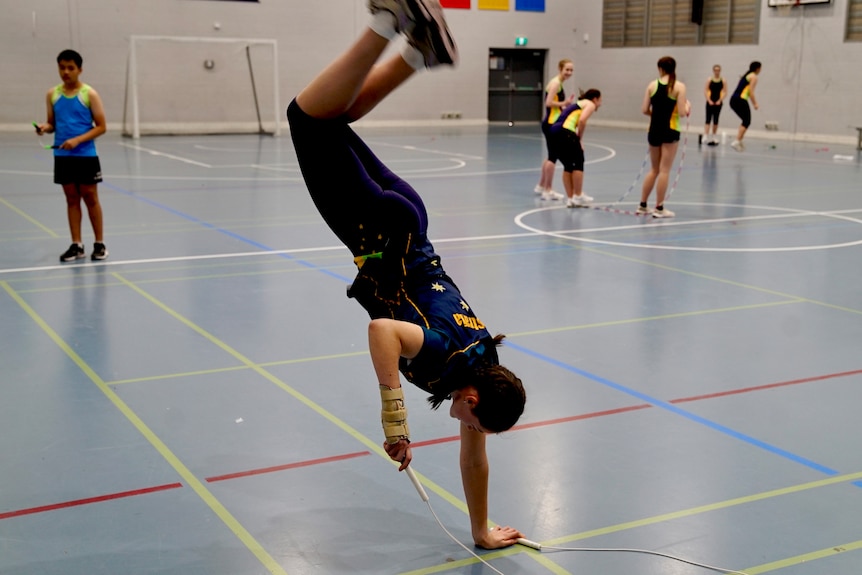 A young woman doing a one-handed handspring while jump roping in a gym.