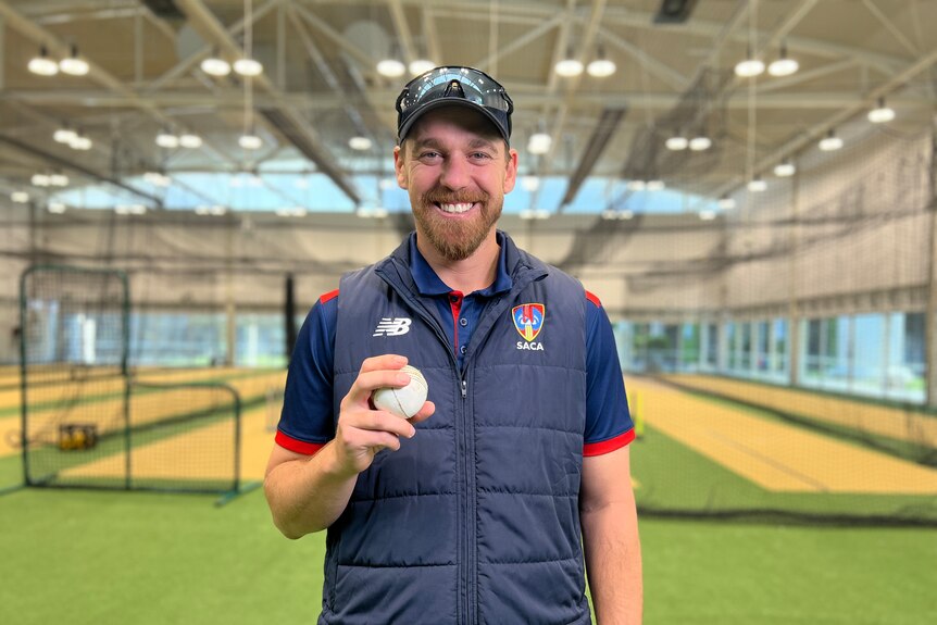 Luke Butterworth smiles as he holds a white cricket ball in his hand