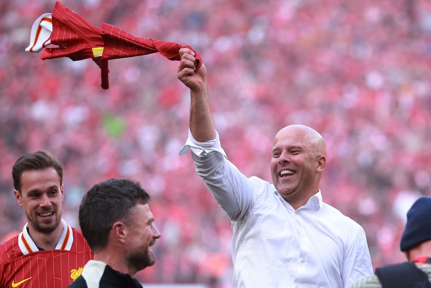 Arne Slot smiling and spinning a Liverpool jersey above his head in celebration