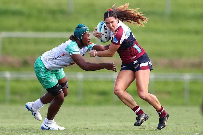Two women, one in green and one in red and white, tussle for a rugby ball in front of greenery.
