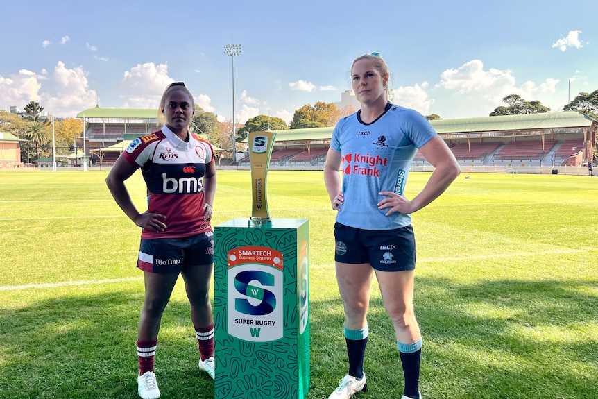 Two women, one wearing a red rugby shirt and the other a blue one, stand either side of a trophy on the side of a field.