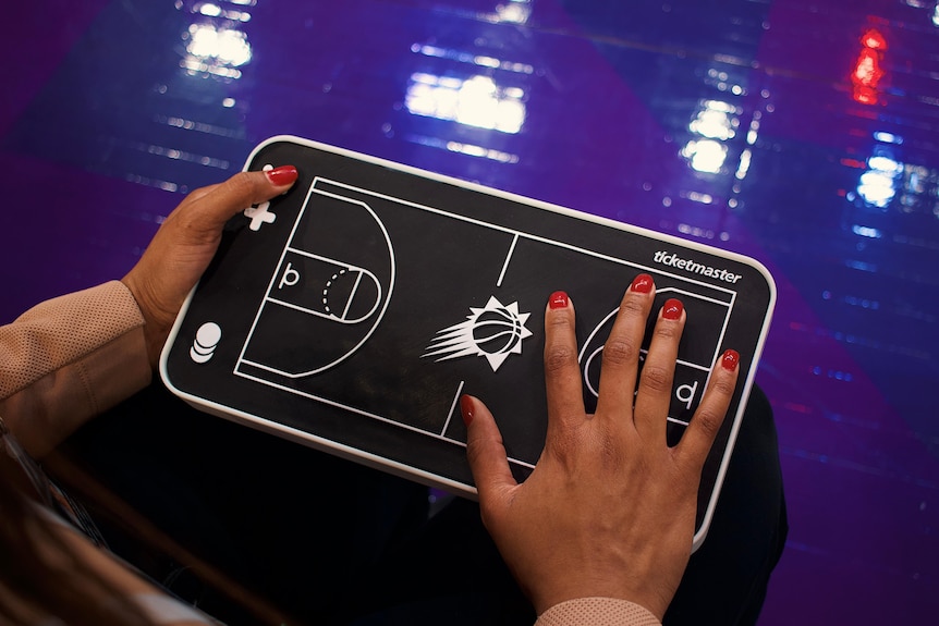 A black, flat model basketball field is being held by one hand and another hand is on its surface with bright red nail polish.