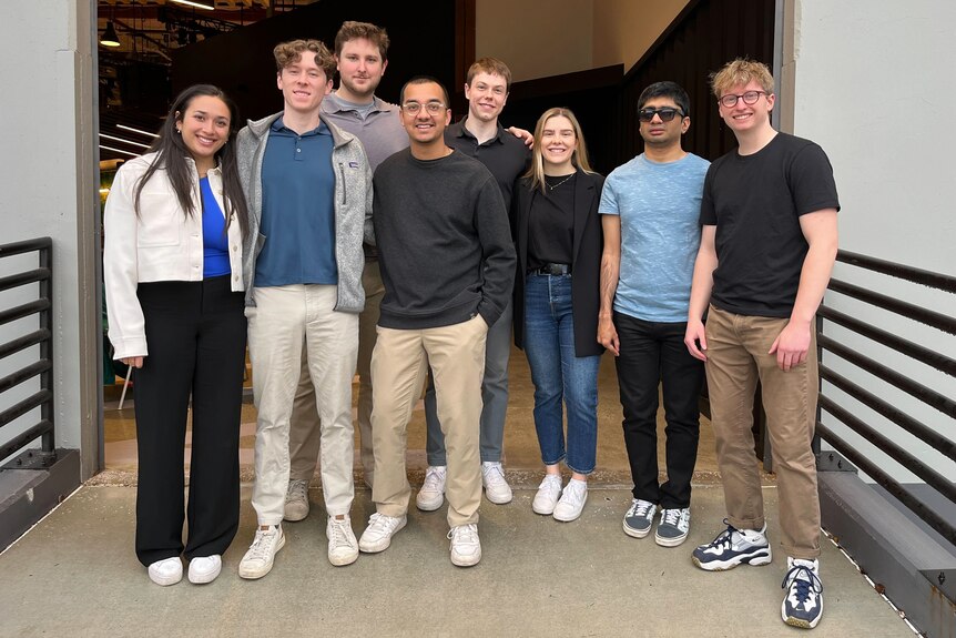 A group of eight mid-20s people pose and smile for a photo in a door way.