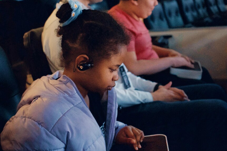 A young girl has her hands on a handheld device and looking down on it, headphone in ear.