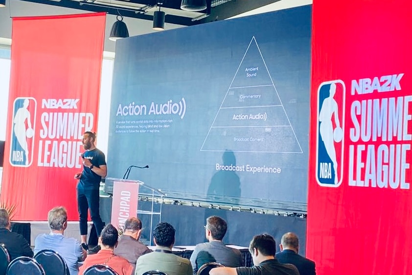 A man in a blue t-shirt and jeans speaks to a crowd in front of an NBA banner and Action Audio graphic.