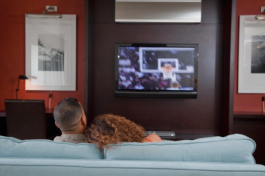 Two people sit on a couch, facing a television showing a basketball hoop and ball going through it.