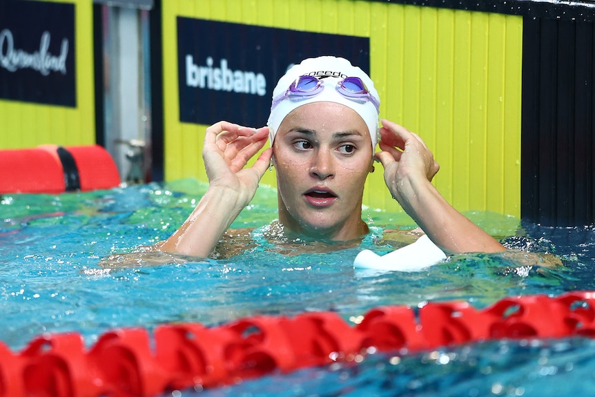 A female swimmer adjusts her cap as she kneels at the end of an Olympic-sized pool.