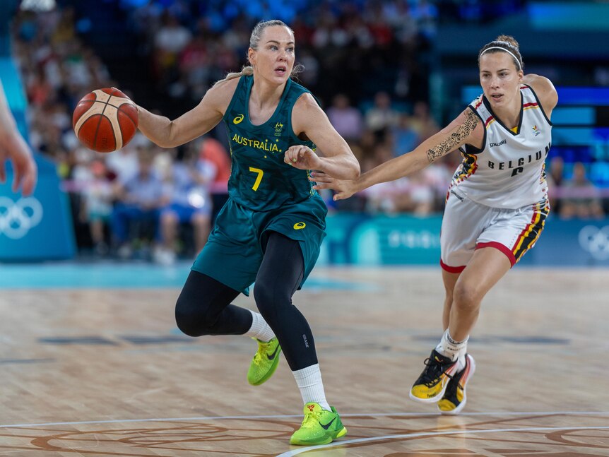 A tall Australian female basketballer uses one arm to fend off a Belgian defender while holding the ball in the other.