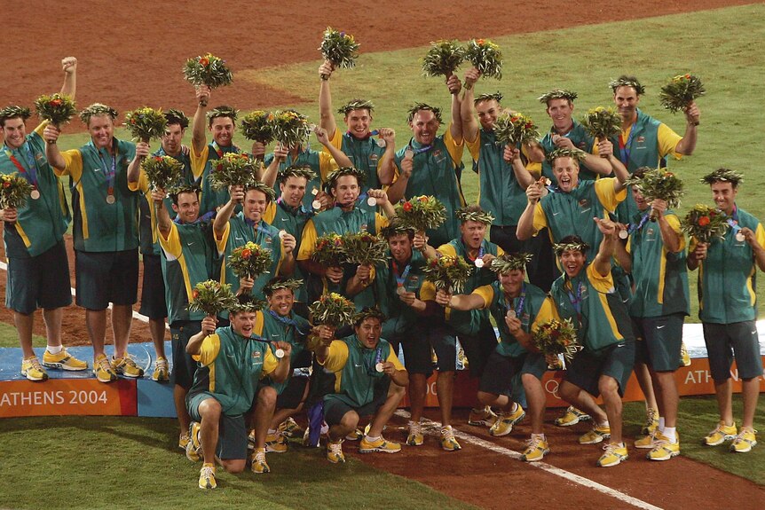 Members of the Australian men's baseball team on the podium with their medals and wreaths on their head, smiling.