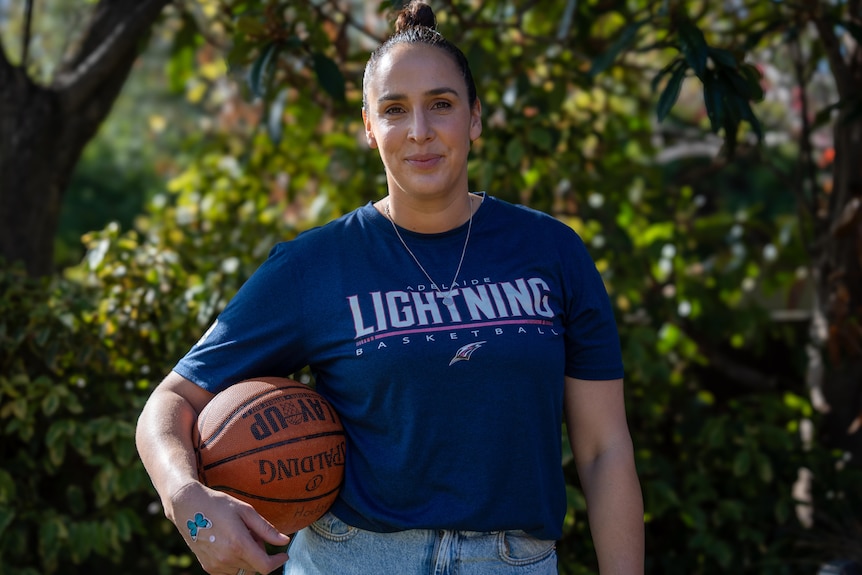 A female smiles while holding a basketball by her right side, wearing a blue shirt says ADELAIDE LIGHTNING BASKETBALL