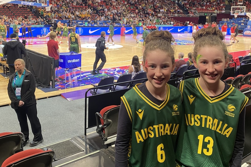 Two twin girls in green and gold uniforms stand in front of a basketball court inside a stadium.