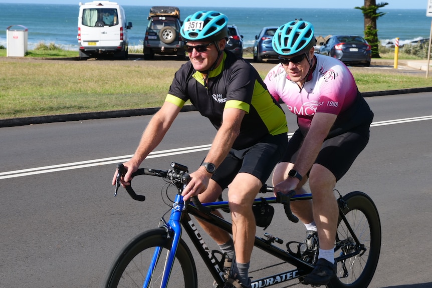 Two men on a tandem bicycle ride along the road wearing blue helmets, with the ocean behind them.