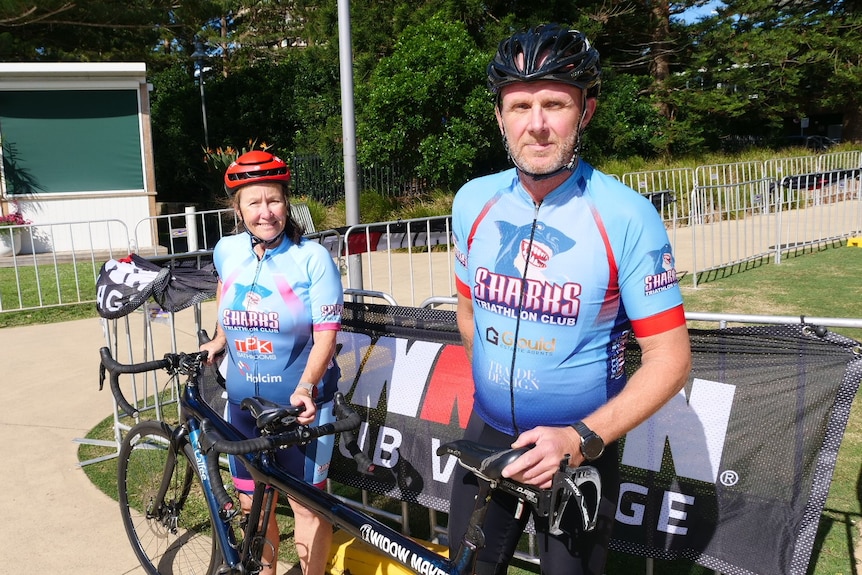 Couple in their fifties standing in lycra and helmets in front of a tandem bike with Iron Man sign on the fence behind them.