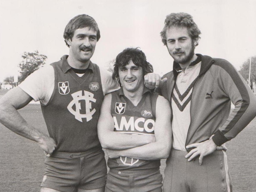 An old photo of two footballers standing with their coach on a football field.
