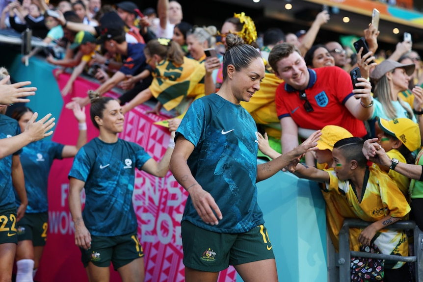 Two soccer players wearing blue shirts and green shorts walk out of a tunnel with fans wearing yellow behind them