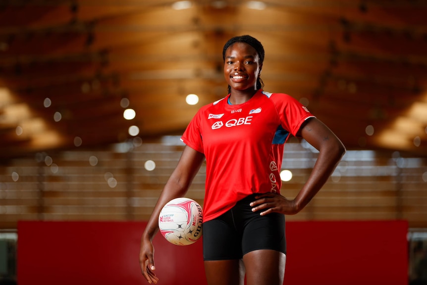 A player wears a red top and stands with their left hand on their left hip as their right arm balances a ball on their right hip