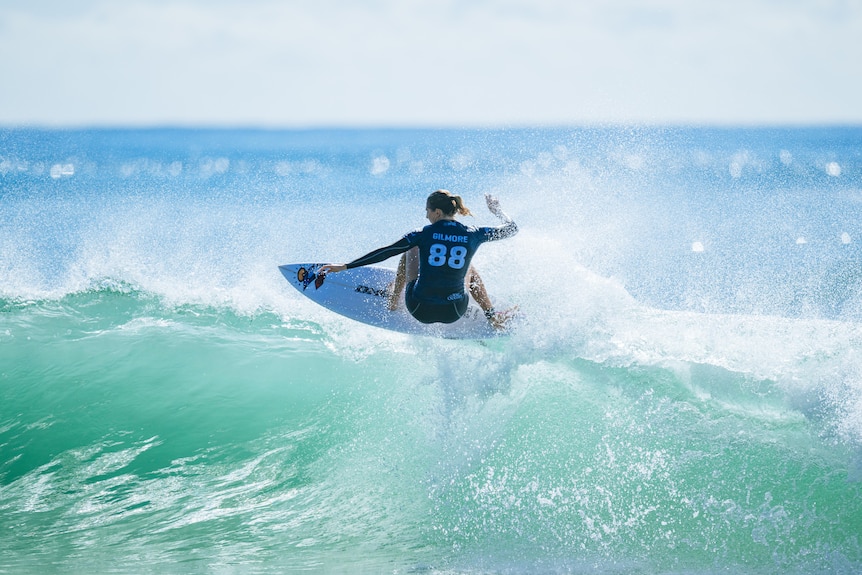 Steph gilmore effortlessly floats on top of a breaking wave, her back to the camera.