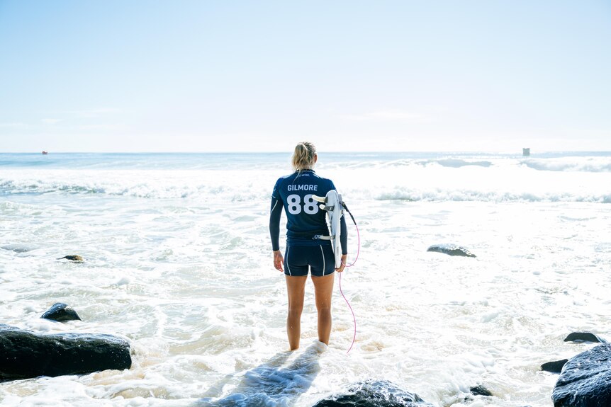 Steph gilmore standing on a rock looking out at the ocean holding her surfboard.