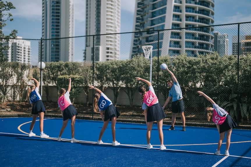 Young teens bend over to their left and warm up for a game while wearing netball attire