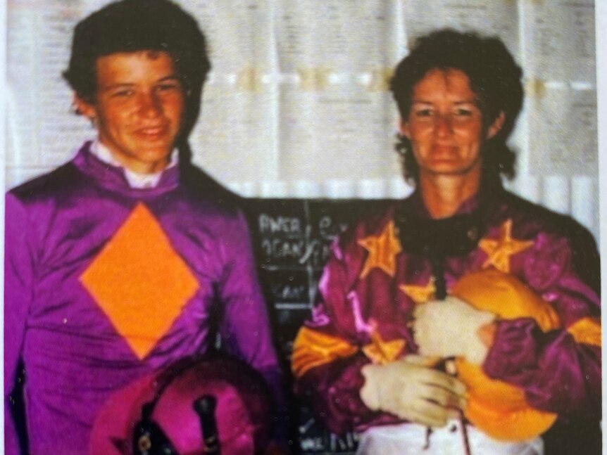 Woman and young man dressed in horse racing silks and holding riding helmet and whip, smiling. 