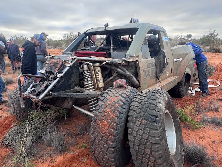 Spectators stand around a wrecked racing truck in the desert.