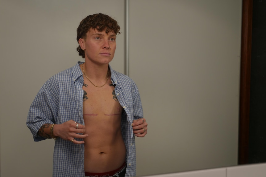 A person looks at themselves in the mirror with the shirt unbuttoned.