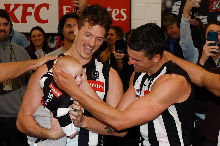 Men in black-and-white striped jerseys hold a baby in a similar jersey and look excited.