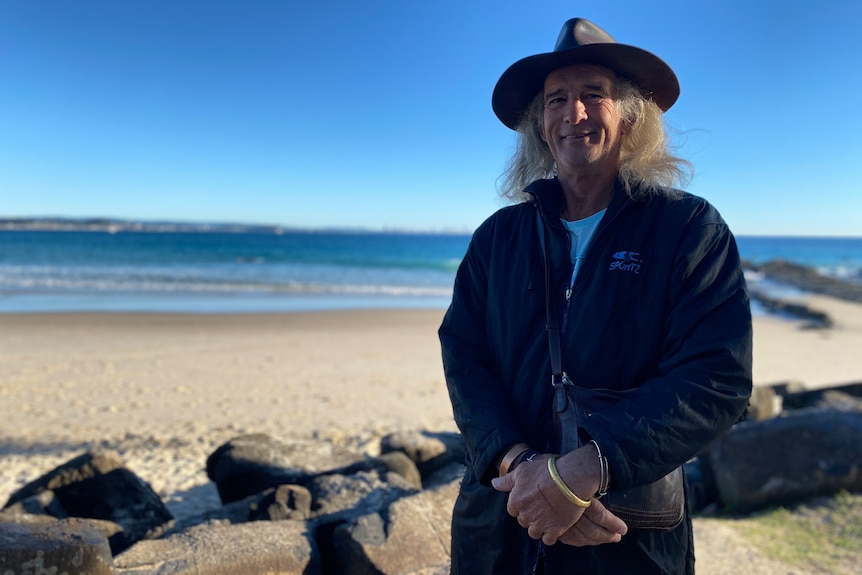 A man in a long black coat and a wide-brimmed hat stands in front of the sand and beach at Snapper Rocks.