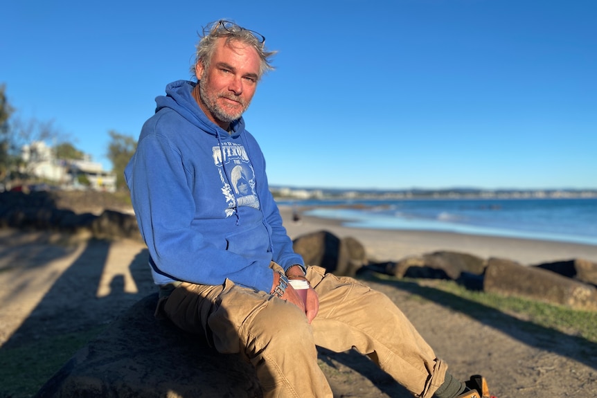 A man in a hoodie and long cargo pants holding a coffee cup while sitting on a rock at the beach.