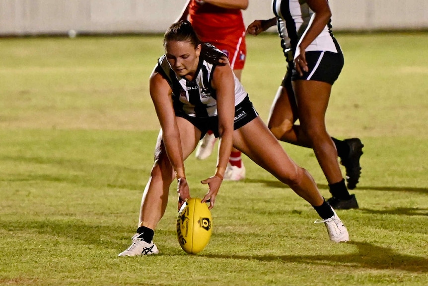 A woman playing afl and she is leaning down to pick a ball up from the ground.
