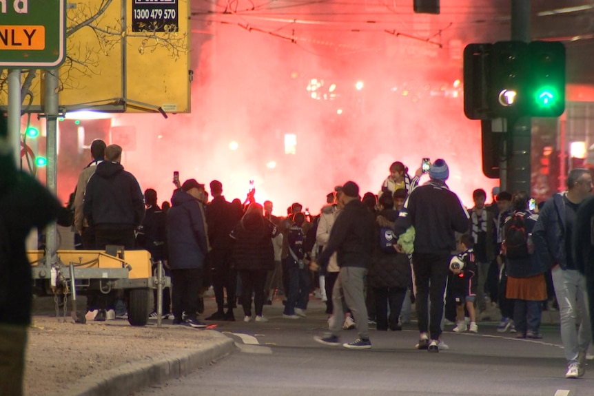 Orange smoke fills the sky behind a crowd gathered on the street at night.