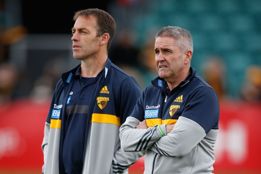 Two men in football branded jackets look out of frame while standing on a football ground.