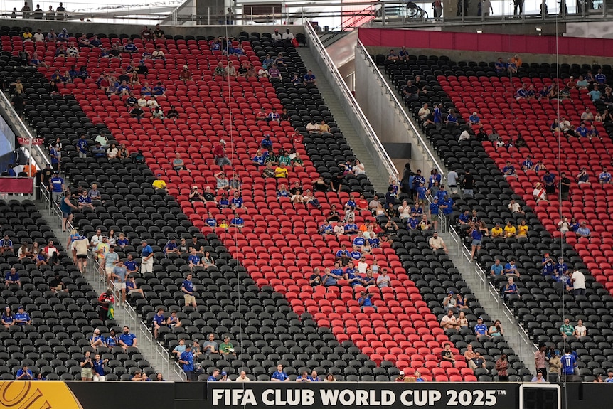 A smattering of football fans in blue jerseys sitting amid rows of empty red and black stadium seating