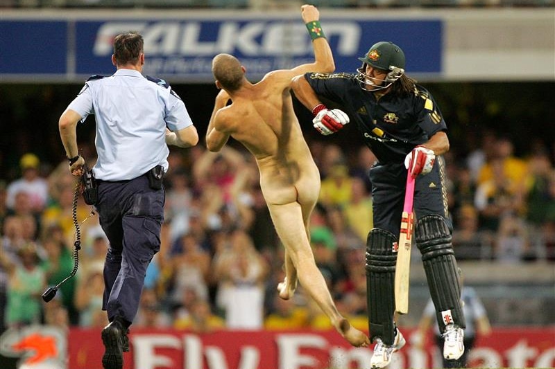 A naked man is knocked over by a man wearing cricket attire and holding a cricket bat