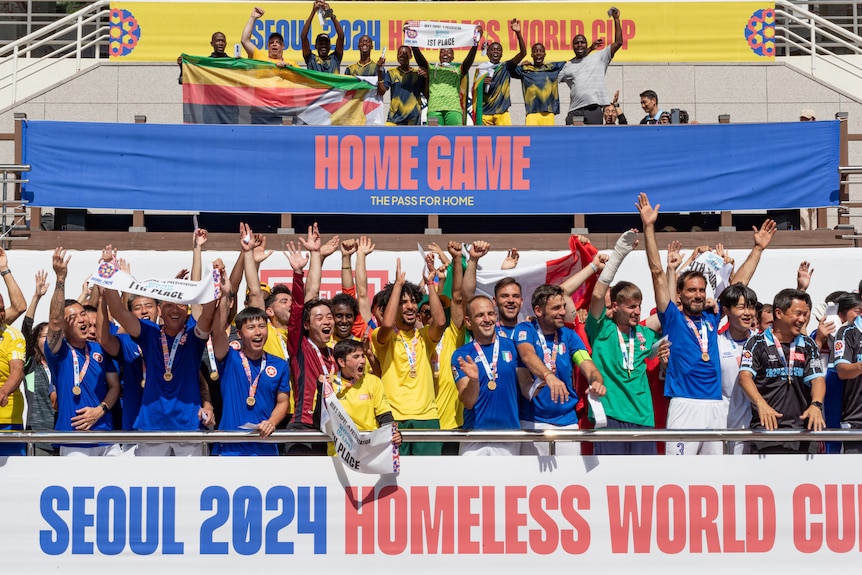 A photo of soccer supporters standing in front of the Seoul 2024 Homeless World Cup banner. It's colourful and exciting.