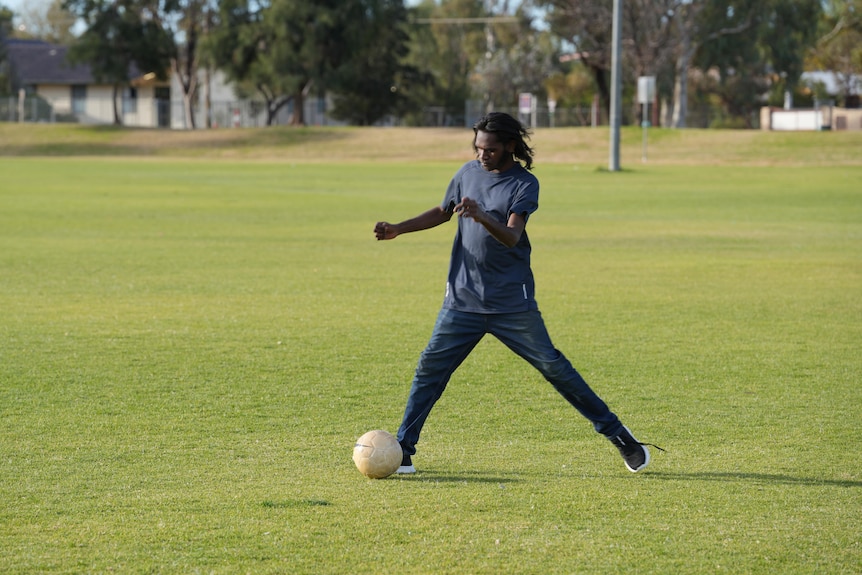 A man kicks a soccer ball on a green oval. His legs are wide in motion.