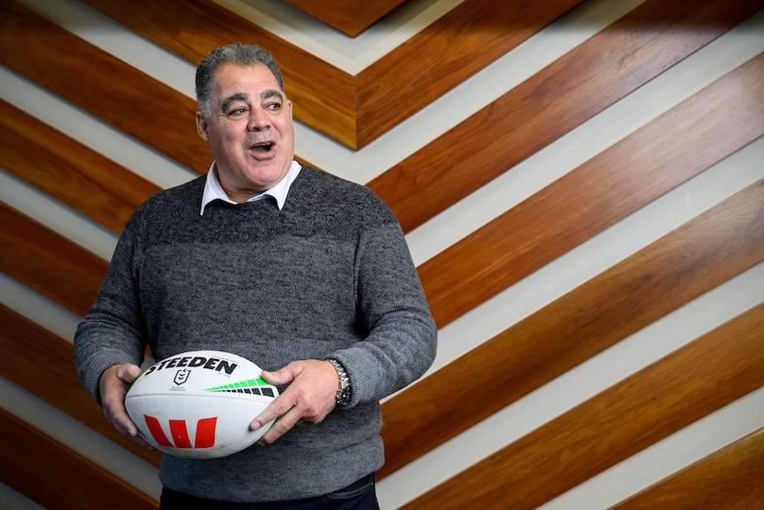 A man in a grey woollen jumper, holding a white football and smiling.