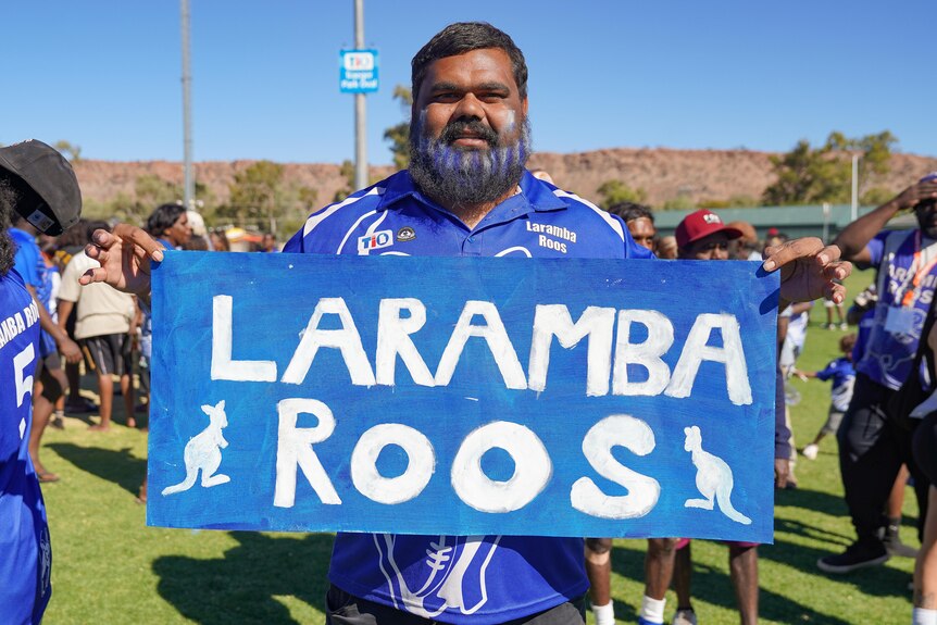 A man with blue and white hair spray on his beard, holding a home-made sign that reads: Laramba Roos.