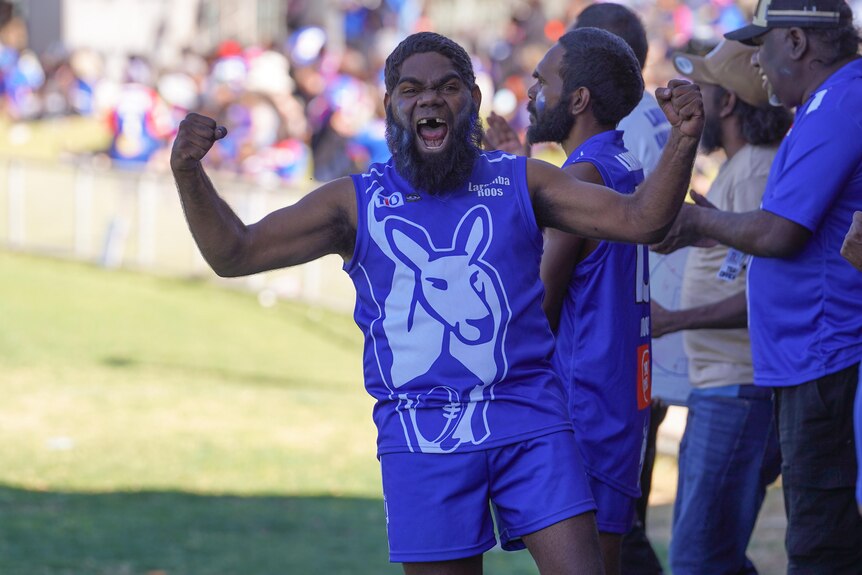 A man wearing a Roos football uniform, cheering from the sideline.