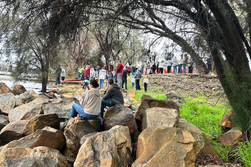 People sit of rocks and stand along a river bank. 