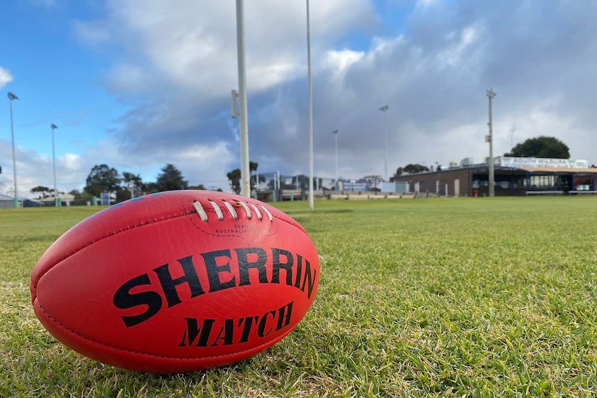 red sherrin football to sitting on green grass with football goal posts and football club in the background