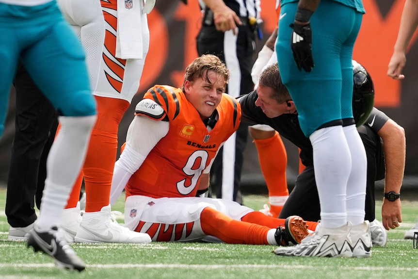 Cincinnati Bengals quarterback Joe Burrow sitting on the ground, speaking to a trainer