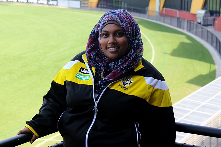 Slightly smiling Rana Hussain, wears a head scarf, a yellow and blue sports jacket and stands in a stadium, footy oval behind.