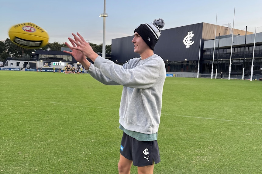 A man in a beanie catches a football on a field with the Carlton logo in the background.