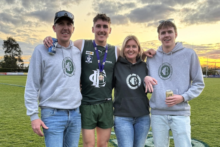 Three men and a woman with arms around each smile at the camera on a grass football field.