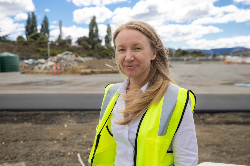 A woman wearing a high vis vest smiles into the camera whilst standing in front of a construciton site.