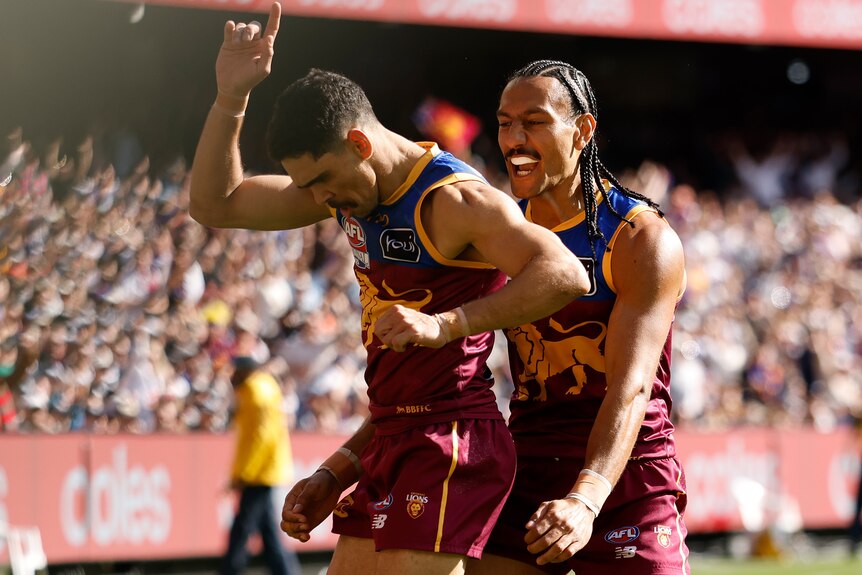 A Brisbane Lions player looks down as he points to the sky in celebration as a smiling teammate stands behind him.