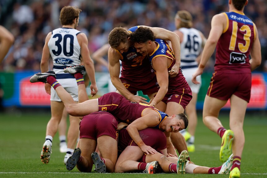 A group of Brisbane Lions players pile on top of each other in celebration on the field, as dejected Geelong players walk away. 