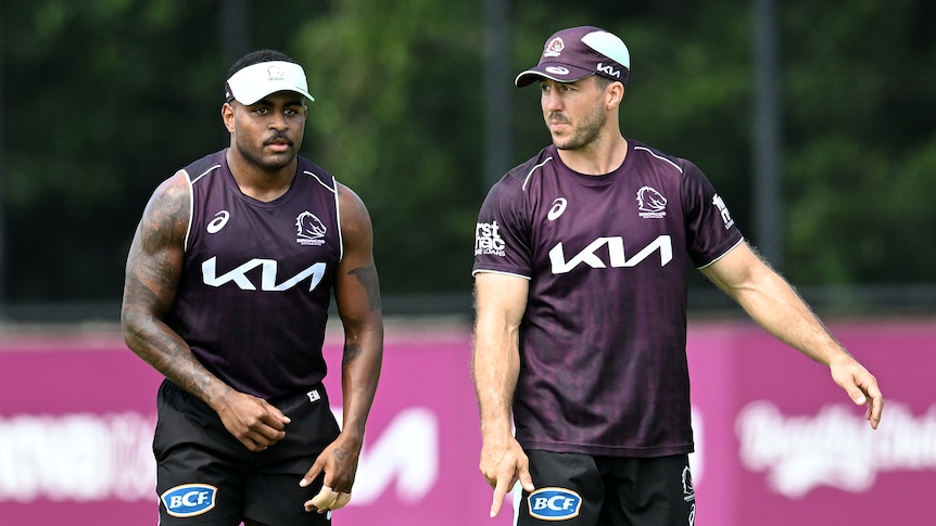 Two Brisbane Broncos players stand next to each other at training as the one on the right looks at his partner.